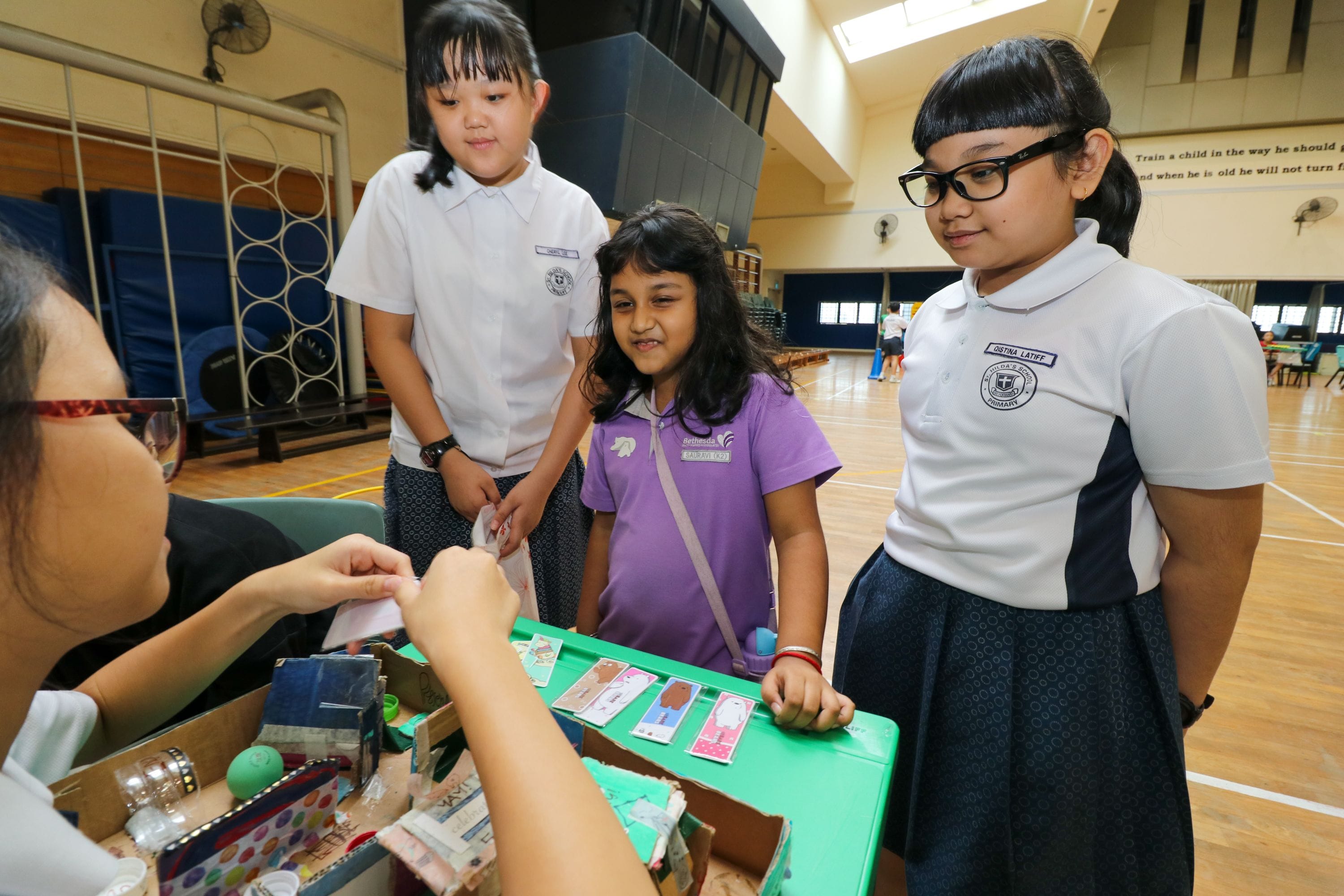 Three schoolgirls in uniforms stand at a table with small items during an indoor school activity event.